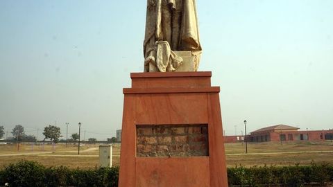 Public art. A panel from a wood base has been removed to expose brick beneath. The base seems to hold up a stone object, perhaps legs wrapped in a cape, but the image is cut off just above the figure’s knees. A grassy lawn leads back to low buildings and power lines in the distance.