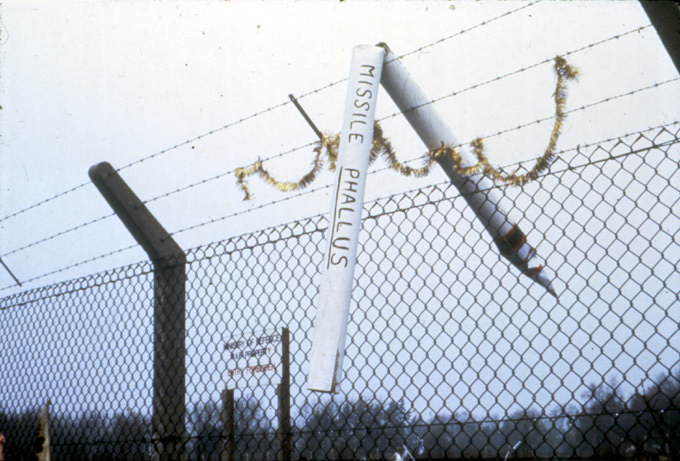 Public art. A long white cylinder with a pointy end has text reading 'Missile Phallus' written on the side. It hangs, bent in half, over barbed wire. Gold tinsel has been strung along the barbed wire below.