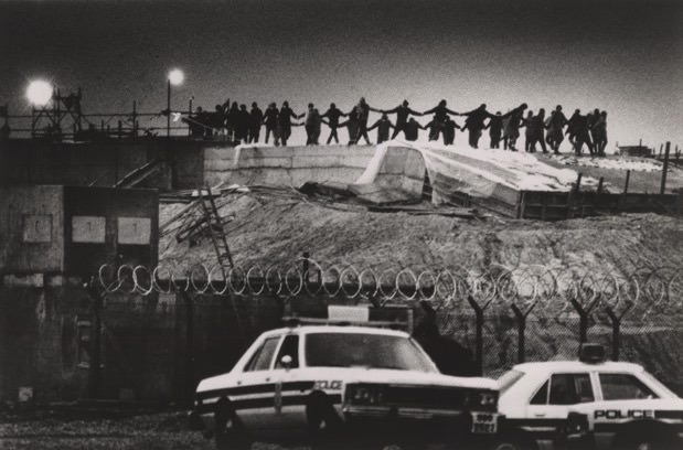 Black and white photograph. A few dozen women dance with hands joined in a ring on a mound. They are black silhouettes against a darkened sky. Two police cars are parked below.