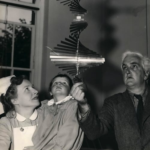 Metal sculpture. Black and white photograph of a senior person holding a metal mobile sculpture and delicately spinning it from its base. A nurse and child gaze up towards the mobile with curious expressions.