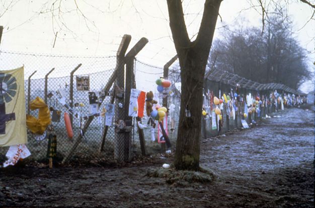 Public art. A chain-link fence topped with barbed wire is hung with balloons, boys, signs, and other colourful objects.