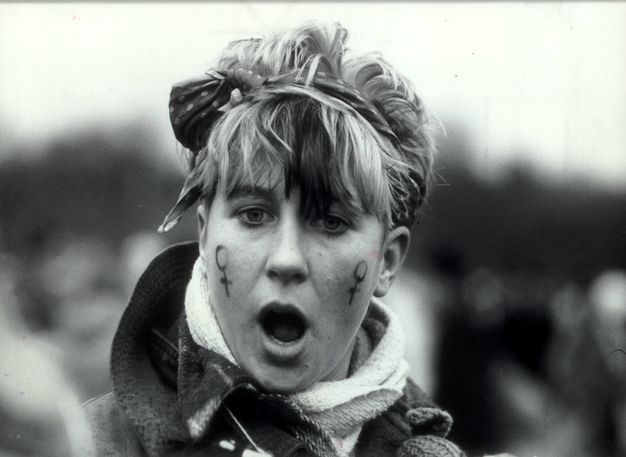Black and white photograph. Shown from the shoulders up, a person with short hair tied behind a scarf faces us, mouth wide open. Two symbols denoting female are drawn on the cheeks.
