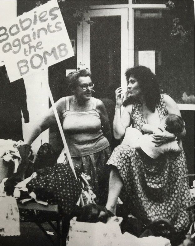 Black and white photograph. A woman holds and nurses a baby. Another woman sits next to her. A protest sign misspells 'against', flipping the t and s.