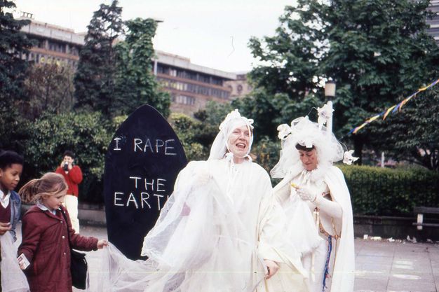 Two pale-skinned women wearing white dresses and gobs of veils tied to their heads stand on a sidewalk in front of trees and a building. One looks up and smiles as she speaks. An arched blackboard behind her reads, 'I Rape the Earth' in white.