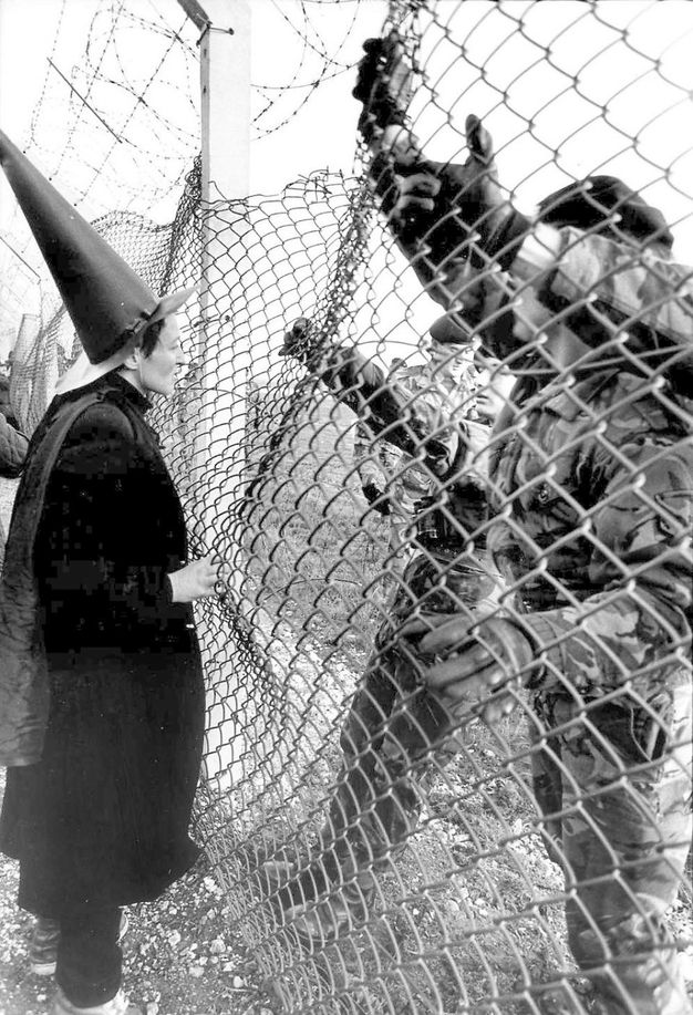 Black and white photograph. A woman dressed in black and wearing a brimmed, conical black hat talks across a chain-link fence with at least four men in camouflage.