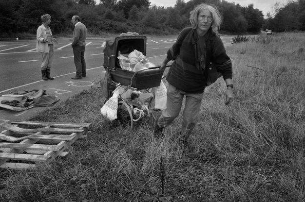Black and white photograph. A woman drags a pram filled and laden with bags through tall grass, away from a road. Two people stand talking in the shoulder of the road.