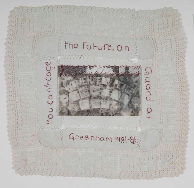 Textile. A black and white image reading 'Women for Peace' is stitched to a square piece of lace. Text also stitched in red around the image reads 'You can't cage the future. On Guard at Greenham 1981-86.