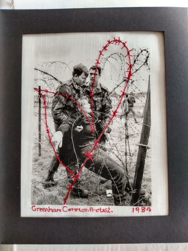 Textile. In black and white, a woman is dragged by two men wearing camouflage uniforms through a chain-link fence topped with coils of barbed wire. Red stitching resembling the barbed wire sweeps across the picture. Text also stitched in red below reads, 'Greenham Common Protest 1984'.