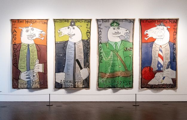 Paintings. Four unstretched canvas panels hang on a white gallery wall. In each, the face of a horse is overlaid on a man’s face. Each wears a different style of uniform. The title is painted across the top and down the sides of the panels. Additional text painted on their shirts is too small to read in this image.