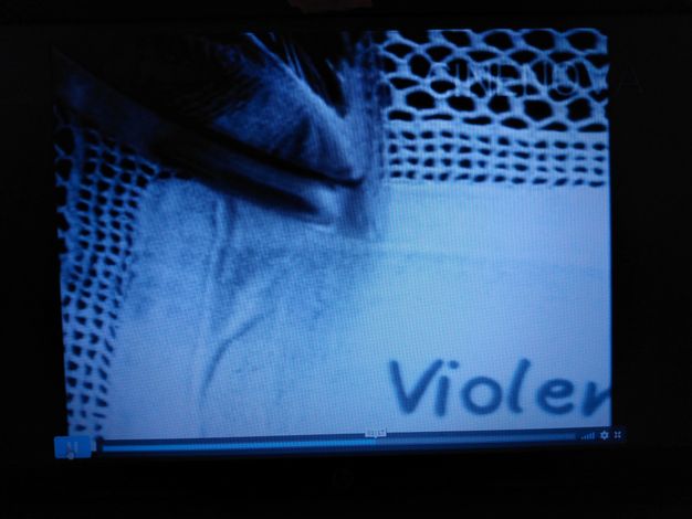 Video still. The pointed tip of an electric iron edges onto the corner of a lace doily. Lettering on the fabric in the lower right corner begins 'Violen' before being cut off by the edges of the frame.