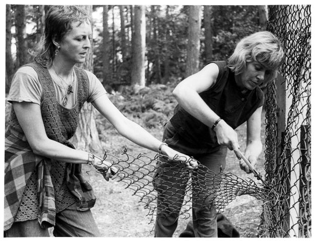 Black and white photograph. One woman cuts through a chain-link fence with wire cutters as another woman pulls the fence away.