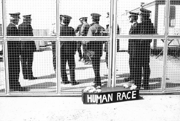 Black and white photograph. A small coffin with the words 'Human race' painted on the side is in front of a closed gate. About nine men in uniform stand behind the fence, all facing away from us.