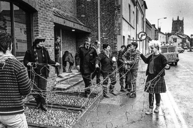 Black and white photograph. Five women hold up the ends of a string web outside a building on the sidewalk next to a street.