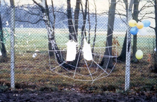 Public art. A web of white string is woven on a chain-link fence. Two white objects on the web are difficult to make out but could be children’s clothing.