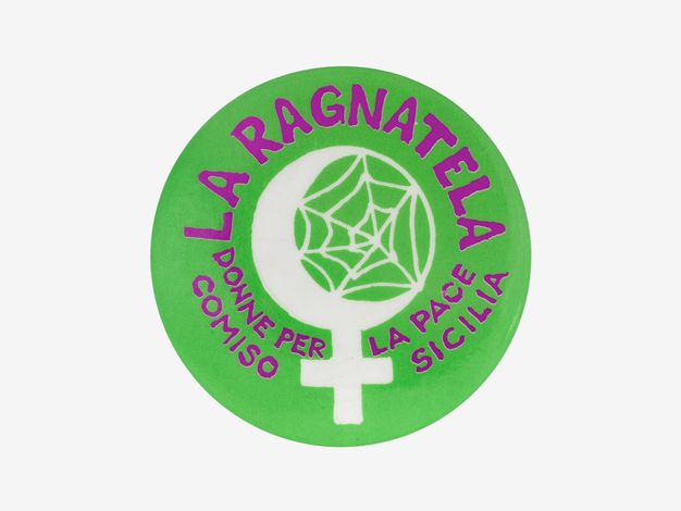 Badge. A green disc has the title in purple letters. At the centre, white a crescent moon holds a spider web. A cross extending from the bottom makes it into the symbol for female.