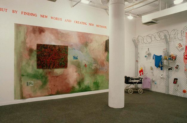 Installation. A mural painting hangs on a wall beneath red text. A section of chain-link fencing topped with coiling barbed wire is to our right. A baby pram sits in front of the fence, which is hung with colourful objects.