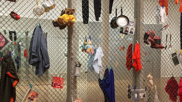 Installation. Detail view of a section of a chain link fence in a gallery is hung with clothing, toys, a frying pan, and other objects.