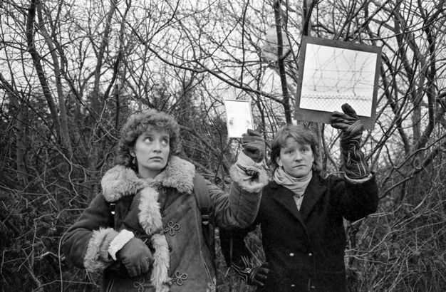 Black and white photograph. Two people wearing coats and gloves hold up pictures in front of a tangle of leafless trees. Light glares off the glass of one picture. The other shows barbed wire.