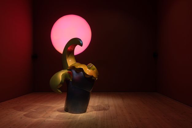Installation. A sculpture has smooth surfaces in midnight blue and golden sage. A columnar base supports a ruffled line, wing-like projection, and a curving hook. A carnation-pink disc glows behind it. The gallery is otherwise dim except for spotlights shining down on the sculpture.