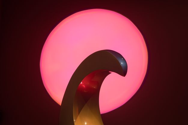 Sculpture. The smooth surface of a curving hood is darkly iridescent against a round pink light.