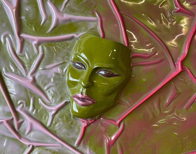 Sculpture. A shiny moss-green mask with high cheekbones, dark eyes, and glittering pink lips appears to sit against a green background veined with lilac purple and hot pink.
