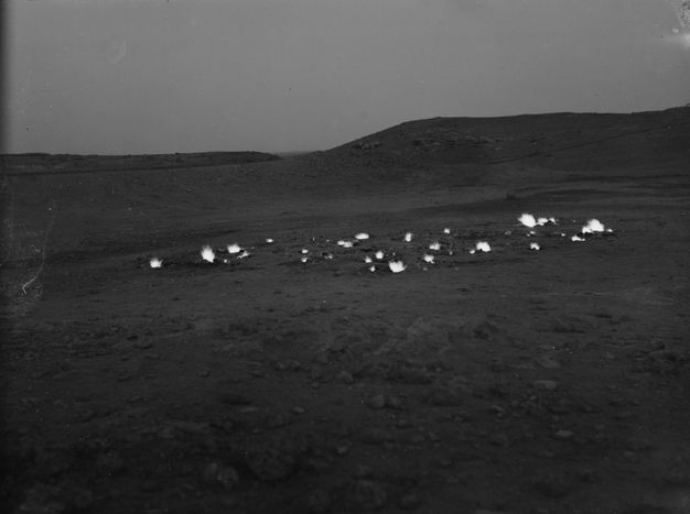 Black and white photograph. Bursts of white flame flare across a barren, rocky landscape.