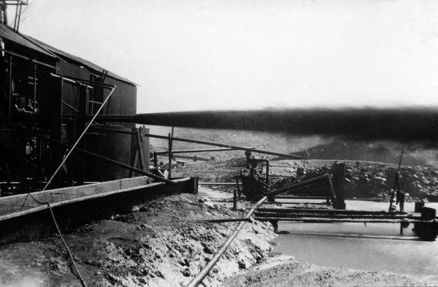 Black and white photograph. Dark oil shoots from a nozzle emerging from a boxy structure to our left. The land around the housing and pipes surrounding it is rocky and treeless.