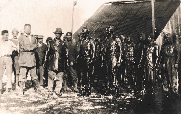 Black and white photograph. Nineteen men stand in front of a structure with s steeply pitched roof. Some of the six standing to our left smile. The rest are covered completely in slick black oil. A pool of oil surrounds their feet to our right.