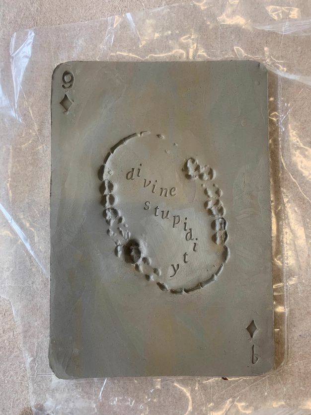 Metalwork. A plate with rounded corners like a playing card has a number nine and a diamond at the top left and bottom right corners. An oval at the centre is a raised impression of a full set of human teeth biting down hard. Lettering inside reads 'divine stupidity'.