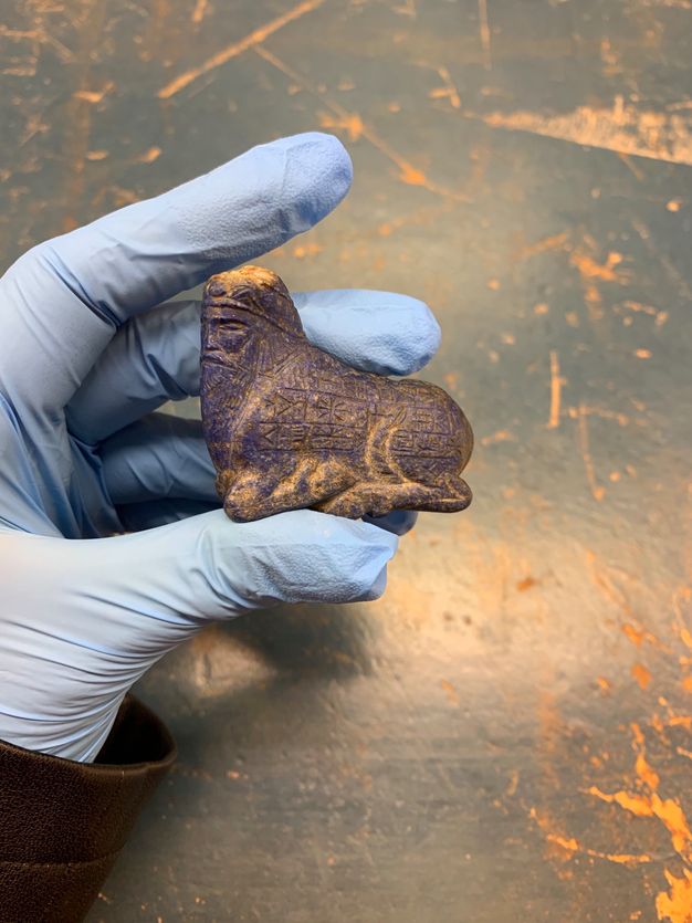 Sculpture. A gloved hand presents a carved object no longer than the pointer finger holding it. The object shows a bull with the head of a bearded man facing our left. Hints of purplish-blue glow from behind a layer of brown.