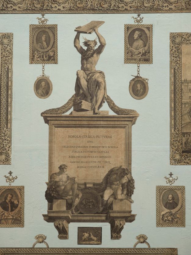 Installation. Detail view of a frontispiece showing a muscular man holding up two tablets. Rays emanate from his wild, curly hair. He sits on a plaque with Latin text that begins, 'Schola Italica Pictvrae'. Two more muscular men below twist or writhe on blocks to either side of a medallion showing a wolf suckling two babies. This frontispiece and other prints are affixed to a pale blue wall.