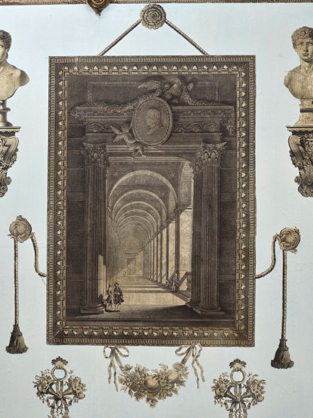 Print. An elaborately framed door with a medallion portrait held up by winged, child-like putti at the top centre opens onto a deep, hall-like with round arches. Two people at the base of the opening are tiny in scale. The print seems to hang from a rosette on a pale blue wall.