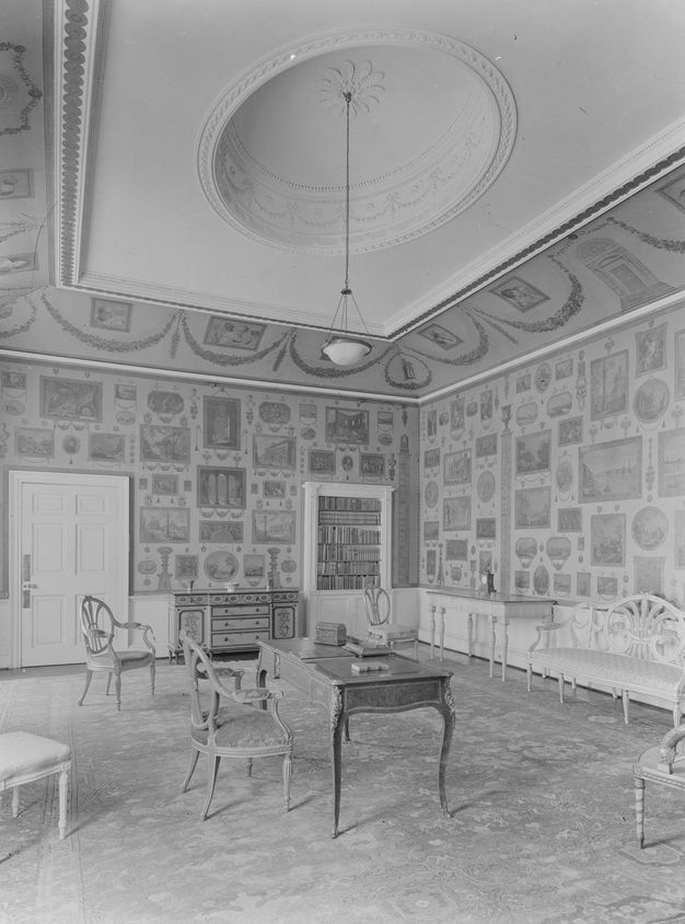 Black and white photograph. A room covered with prints is furnished with wood, oval-backed chairs, a desk, sideboard, and a bench.