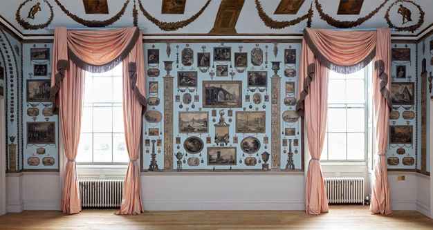 Installation. A pale blue wall has two windows draped with rose pink curtains. Prints are carefully and tidily arranged in the spaces between and around the windows.
