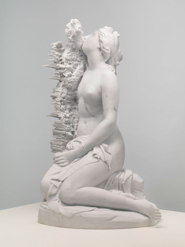 Sculpture. A mostly nude woman kneels with a cloth across her hips. Her hair is plaited, and her face is lifted, facing our left in profile. A form next to her is difficult to make out. It looks like vegetation along her body but turns into long spikes on the far side.