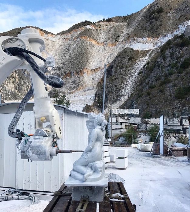 A machine with a needle-like tool carves a slab of marble into the form of a kneeling woman. The machine and other marble slabs are at the foot of steep mountains.
