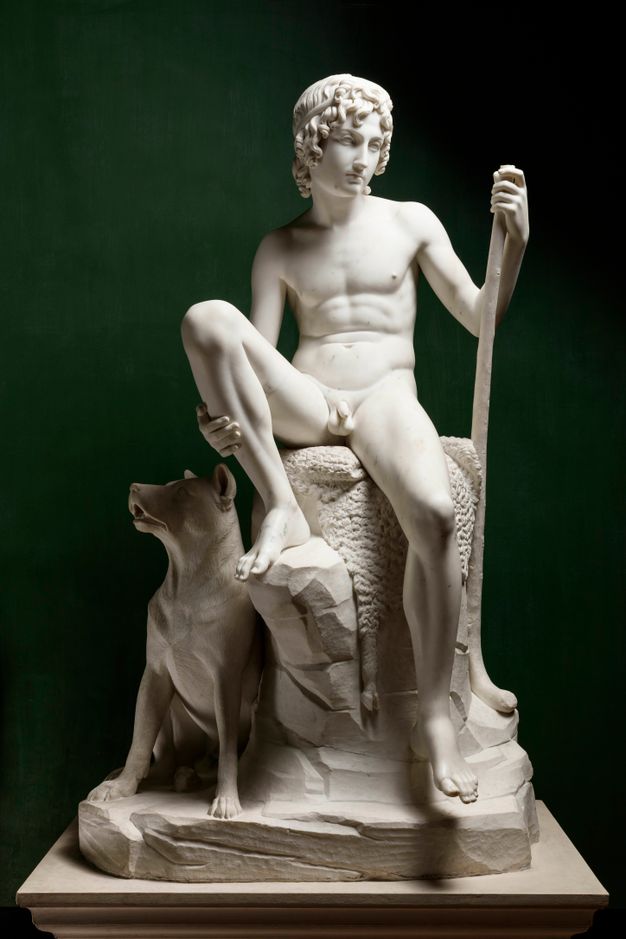 Sculpture. A nude boy sits with one foot propped up on a rock. He holds onto that shin with one hand and the other rests on a walking stick. A dog sits next to the rock.
