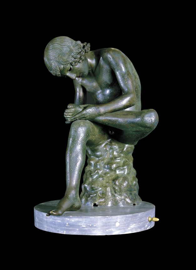 Sculpture. A young boy sits with one foot propped on the other knee. He bends forwards to inspect the underside of that foot, his head bowed. The free-standing sculpture is on a round marble base.