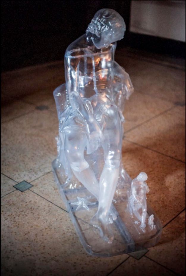 Sculpture. We look slightly down onto the form of a young boy sitting and sleeping, his head bowed forwards. The work is made with clear resin. The whole form is repeated a shin-high version on the base next to the boy’s feet, and then an even smaller, ankle-high version is next to that.