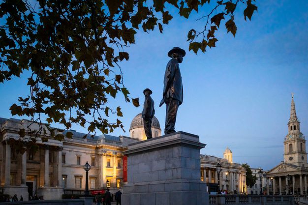 Public art. Two men stand on a high, narrow platform. Seen from below so they are outlined against a sunset blue sky, they face away from each other from each narrow end of the stone base. Buildings enclose the space of this city square.