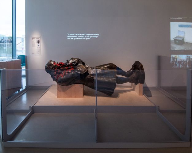 Installation. A bronze sculpture of a standing man is displayed lying across wooden supports in a gallery. The statue has been spray painted with red on the face and across the chest. Text projected onto the wall above it reads, 'Colston’s statue *has* taught me history. When I see it, I reflect on the bad things and feel grateful for the good'.