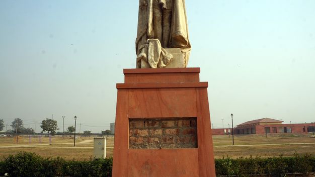 Public art. A panel from a wood base has been removed to expose brick beneath. The base seems to hold up a stone object, perhaps legs wrapped in a cape, but the image is cut off just above the figure’s knees. A grassy lawn leads back to low buildings and power lines in the distance.