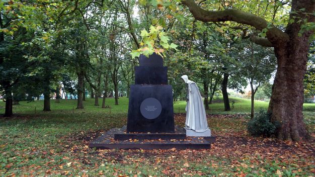 Public art. An empty black plinth is faced with a round plaque. A sculpted person wearing a long robe and white head covering bends to examine something on the plinth. The work is set up under a sprawling tree in a park.