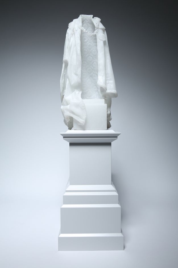 Sculpture. A white cloak hanging as if on a body but without one is on a white plinth. A chain connects the lapels across what should be the chest, and the lining of the cloth is patterned.