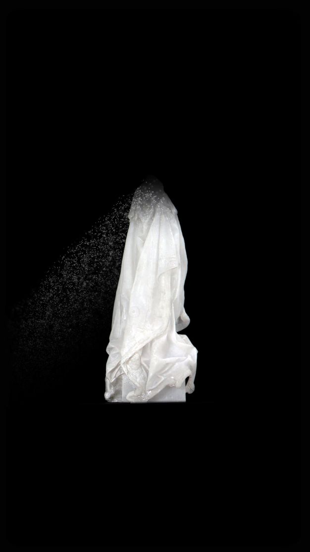 Video still. A white cloak hangs as if over someone’s shoulders against a black background. The top dissolves into white specs and floats down and to our left.