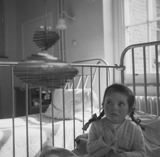 Black and white photograph. Young child in braided pigtails looks up at a spiral-shaped metal mobile sculpture gleaming from the light of a nearby window.