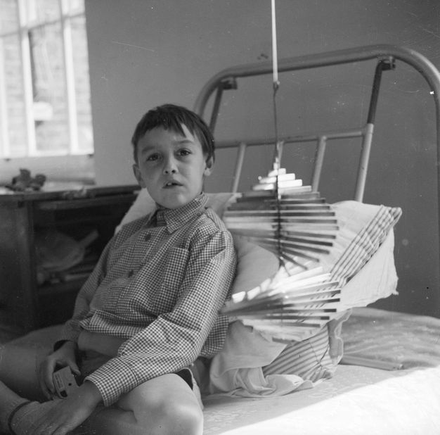 Black and white photograph. Child seated cross-legged on a hospital bed looks towards us with a questioning expression, mouth slightly agape. A metal spiral-shaped mobile sculpture hangs next to the bed. It gradually expands out towards the middle and contracts towards the bottom.