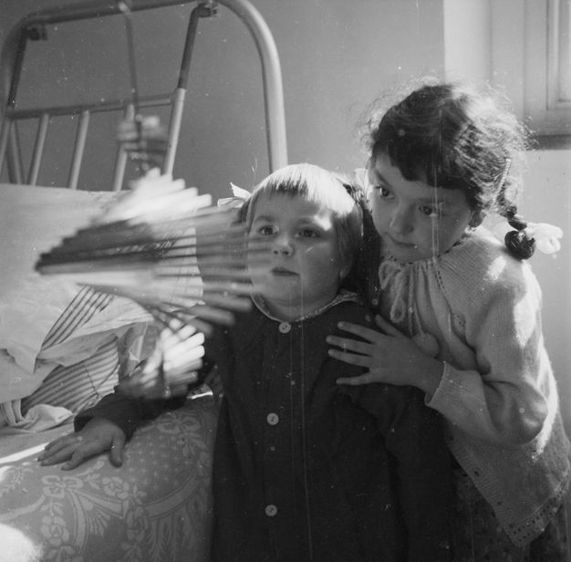 Black and white photograph. Two children stand beside a hospital bed, looking in wonder at a hanging metal spiral mobile sculpture that appears to be spinning and catching the light from the window.