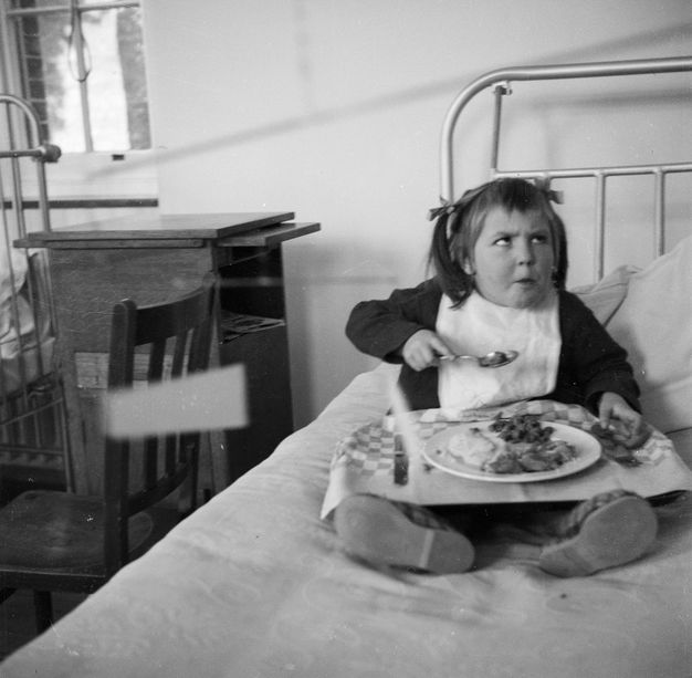 Black and white photograph. Young child seated in a hospital bed, eating from a tray of food on their lap, looking up and furrowing their brow, perhaps with frustration, at something out of frame.