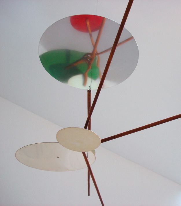 Sculpture. Closeup of the detailing of a mobile with interlocking rods connecting three mirrored circles in various sizes, one reflecting down to reveal additional interlocking rods that connect to green and red circular shapes.